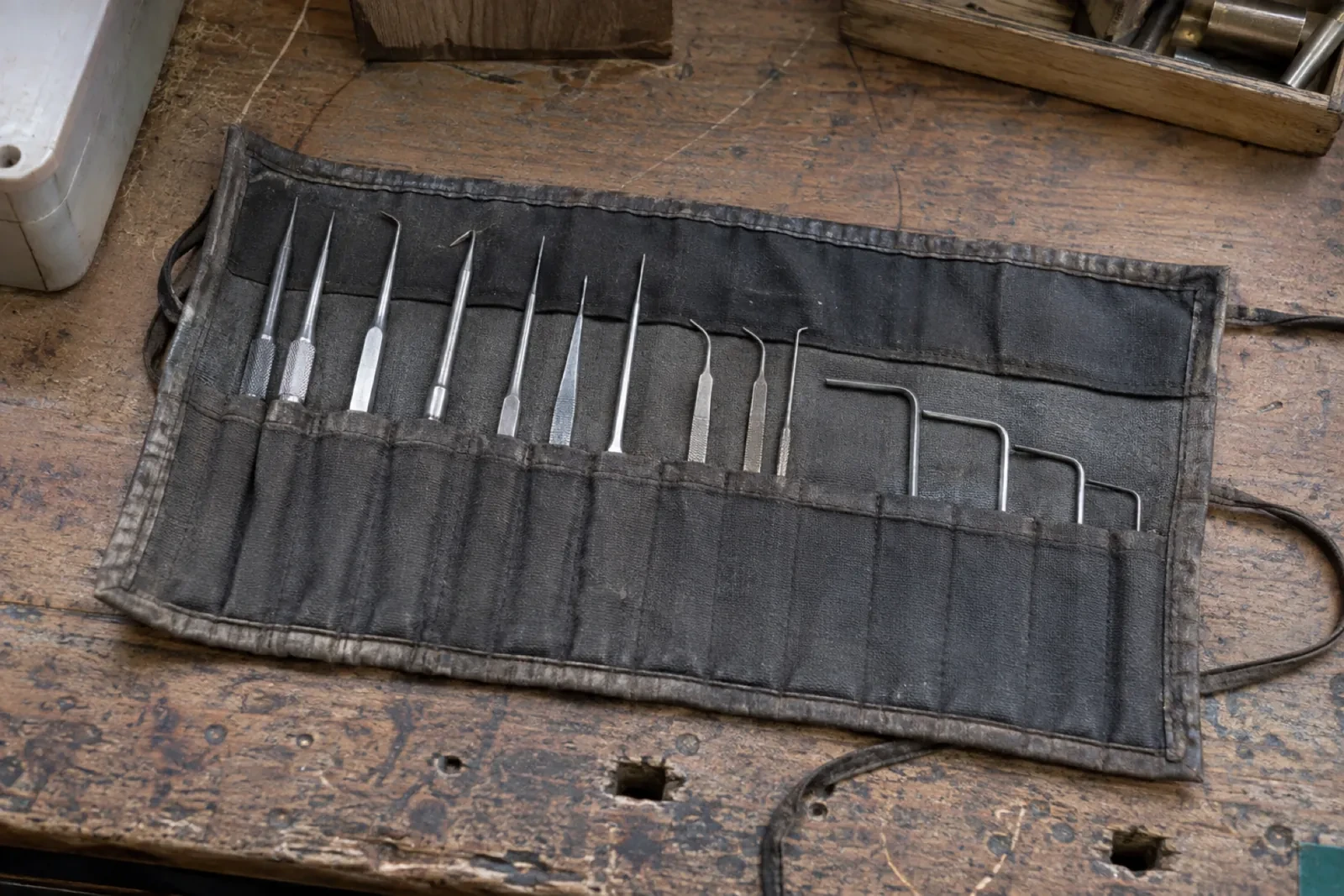 Roll-up black cloth pouch lying open on a worn wooden bench, holding thin metal probes and hooked tools in separate pockets.